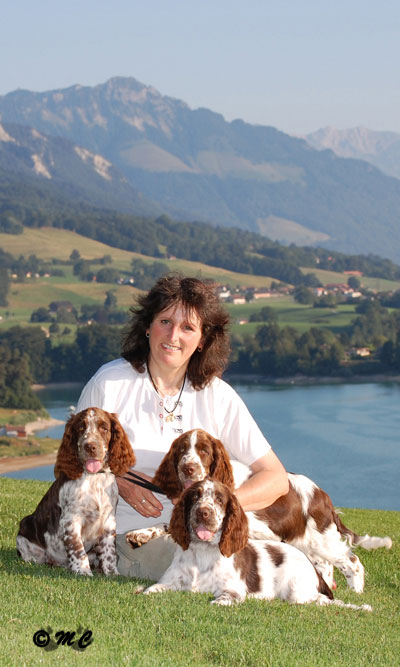 photo de Catherine Thomas assise dans l'herbe du golf de Pont-la-Ville, tenant trois chiots springers spaniels anglais de Syringa, avec le lac de la Gruyère en arrière-plan.