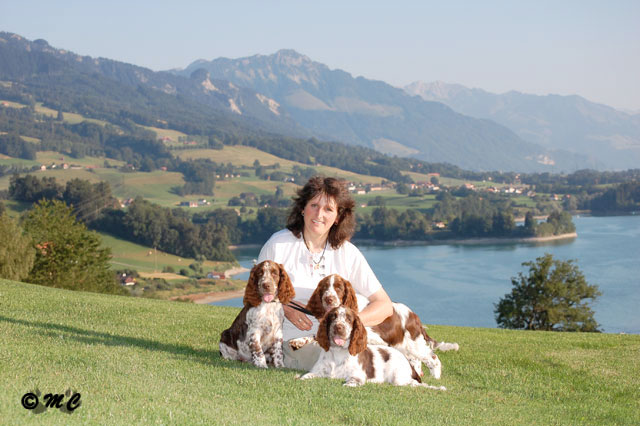 photo de Catherine Thomas assise dans l'herbe du golf de Pont-la-Ville, tenant trois chiots springers spaniels anglais de Syringa, avec le lac de la Gruyère en arrière-plan.